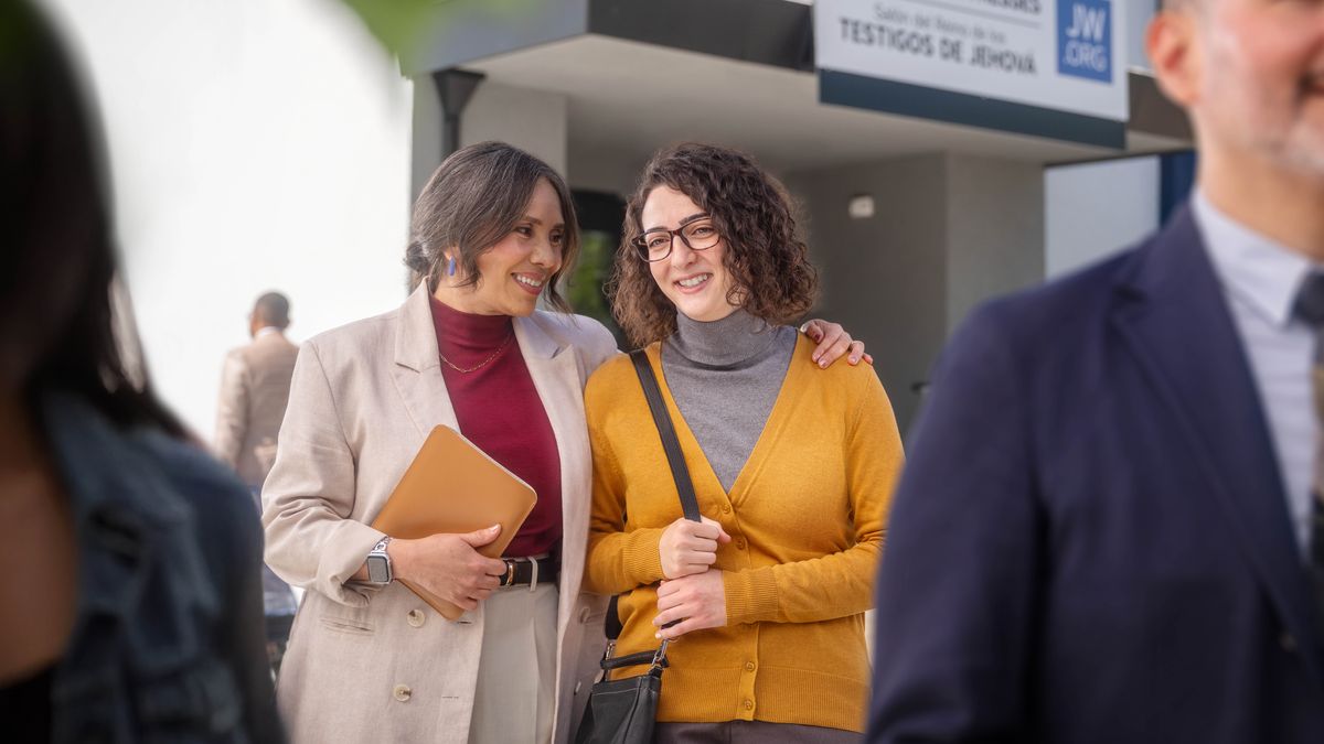 A sister smiling as she leaves a Kingdom Hall to participate in the ministry with another sister who has her arm around her.